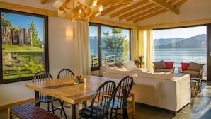 A spacious living area with a wooden dining table, comfortable seating, and large windows offering a view of a lake and mountains. - Correntoso TownHouses (Villa La Angostura)