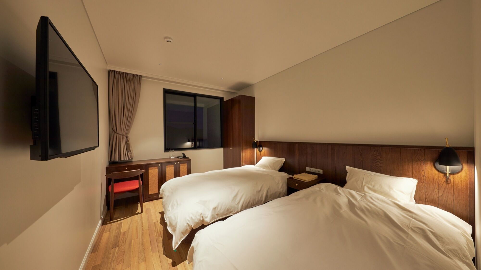 A hotel room with two beds, a desk, a TV, and a window with curtains.