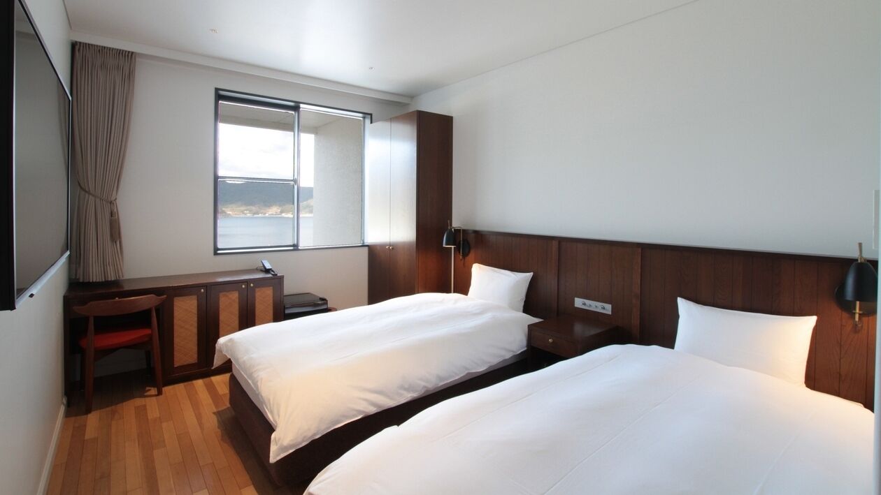 A hotel room with two beds, a desk, a chair, a window with a view, and a wooden headboard.