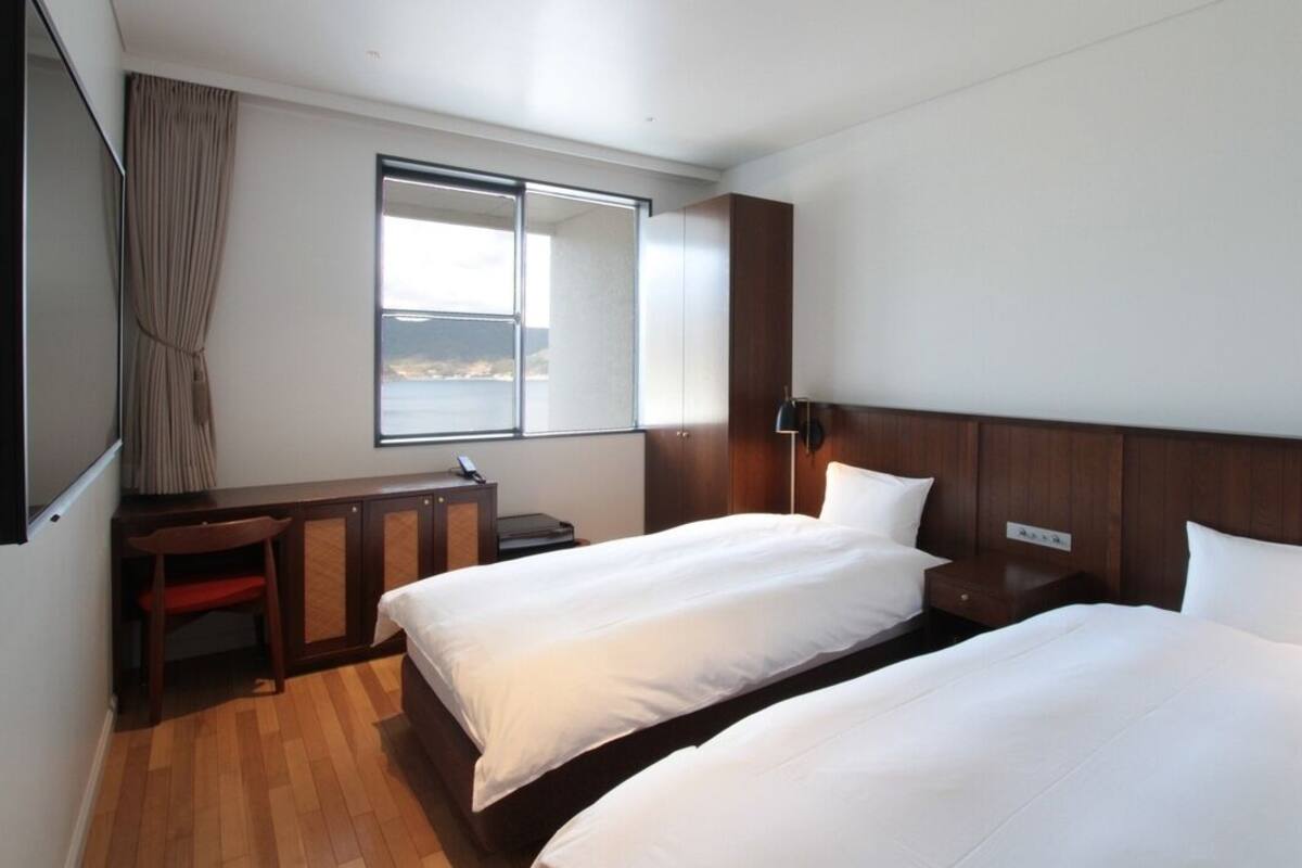 A hotel room with two beds, a desk, a chair, a window with a view, and a wooden headboard.