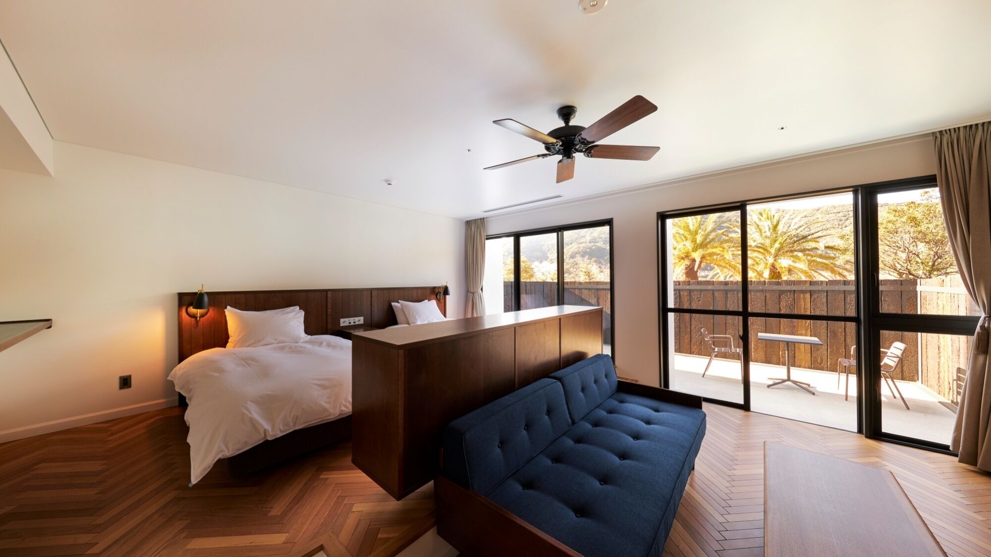 A modern bedroom with a bed, a sofa, a ceiling fan, and large windows.