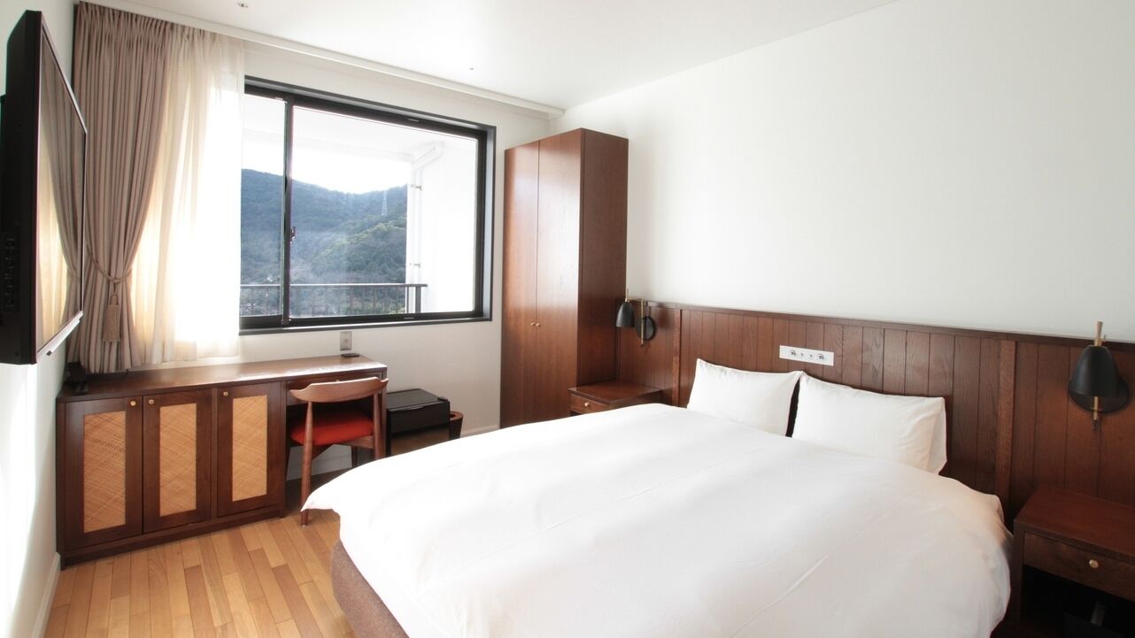 A hotel room with a bed, a desk, a chair, a wardrobe, and a window with a view of the mountains.
