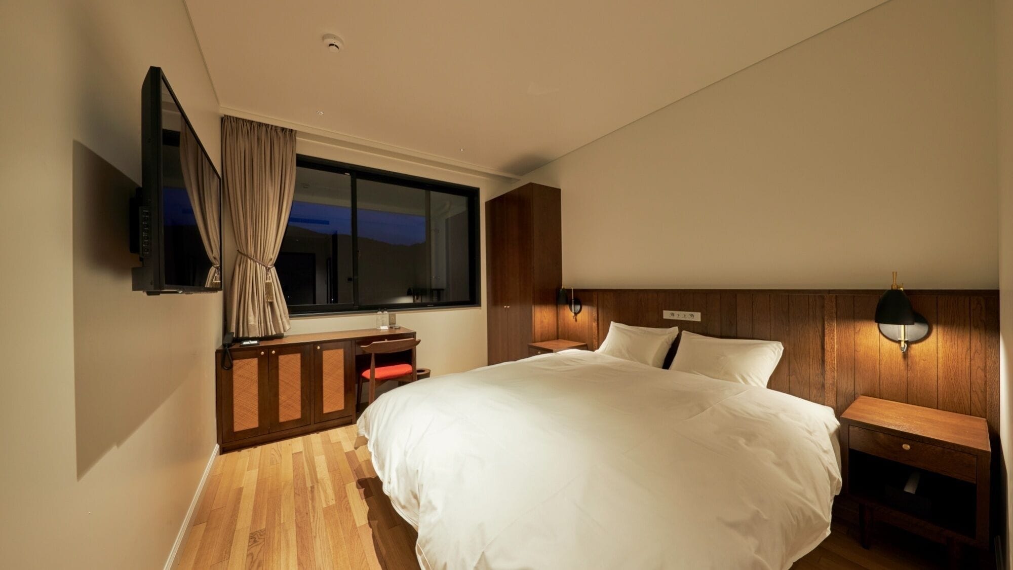 A hotel room with a large bed, a desk with a chair, a TV mounted on the wall, and a window with curtains.