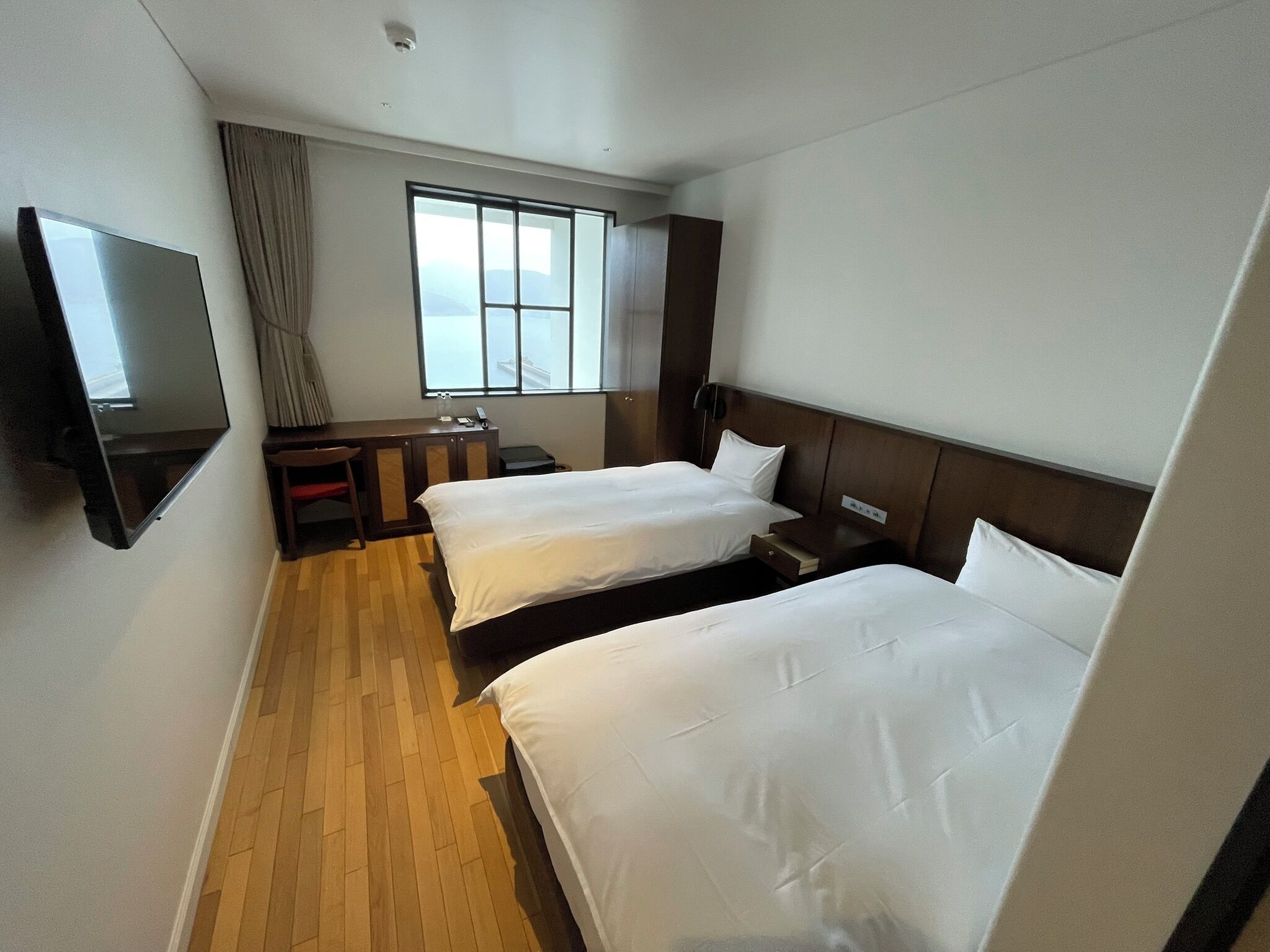 A hotel room with two beds, a TV mounted on the wall, a desk with a chair, and a window with curtains.