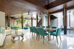 A modern dining area with a large table and teal chairs, a white sofa, and a stone wall with greenery.