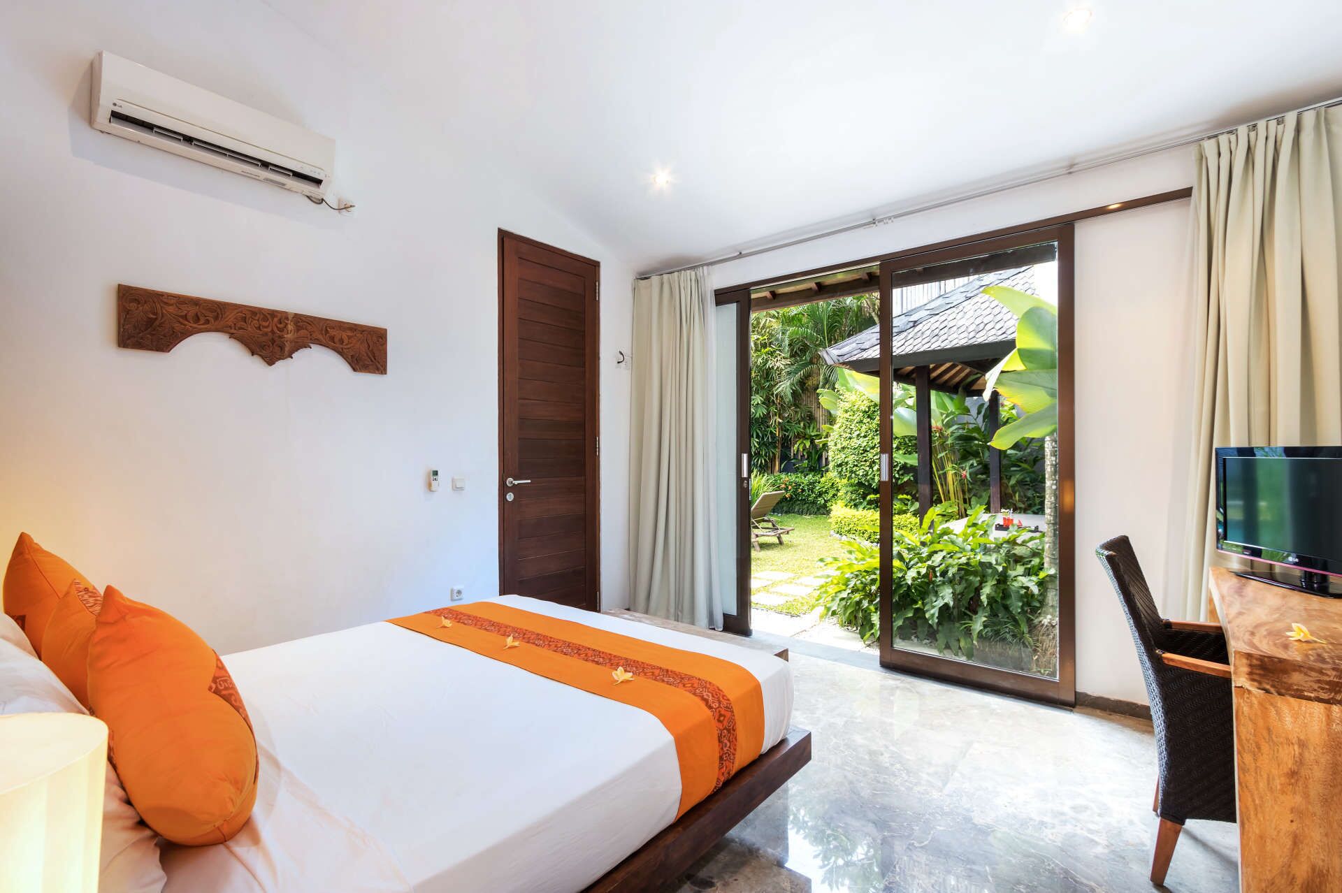 Une chambre d’hôtel avec un lit, un climatiseur, une porte en bois et une vue sur un jardin, accessible par une porte-fenêtre coulissante.