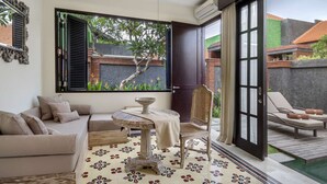 Ein Zimmer mit einer Couch, einem kleinen Tisch und einem Stuhl. Ein Fenster bietet Blick auf einen Garten und einen Poolbereich.