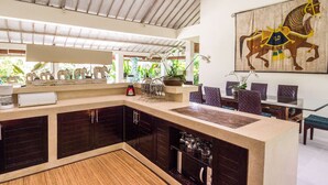A modern kitchen with dark cabinetry, a central island, and a dining area with chairs and a table. - Beautiful Villa With Private Pool, Bali Villa 2051 (Seminyak)