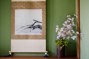 A framed artwork of a bird on a branch, a vase of pink flowers, and a wooden floor.