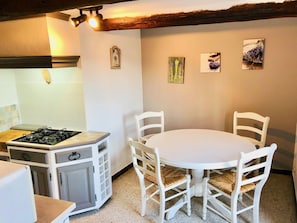 A kitchen with a gas stove, white cabinets, a round table with white chairs, and wall-mounted artwork. - Gîte La Bastide de La Loge (Sault)