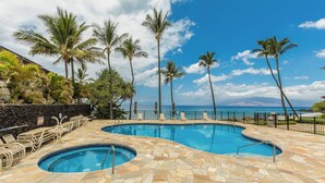 A pool area with two pools, lounge chairs, palm trees, and a view of the ocean. - Beachfront Wailea condo w/ ocean views and lanai (Wailea)
