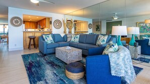 A modern living room with a sectional sofa, a coffee table, and a kitchen area visible through an open door. - Beachfront Wailea condo w/ ocean views and lanai (Wailea)