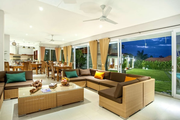A modern living room with a large sectional sofa, a dining area, and a view of a lush garden and pool at night.
