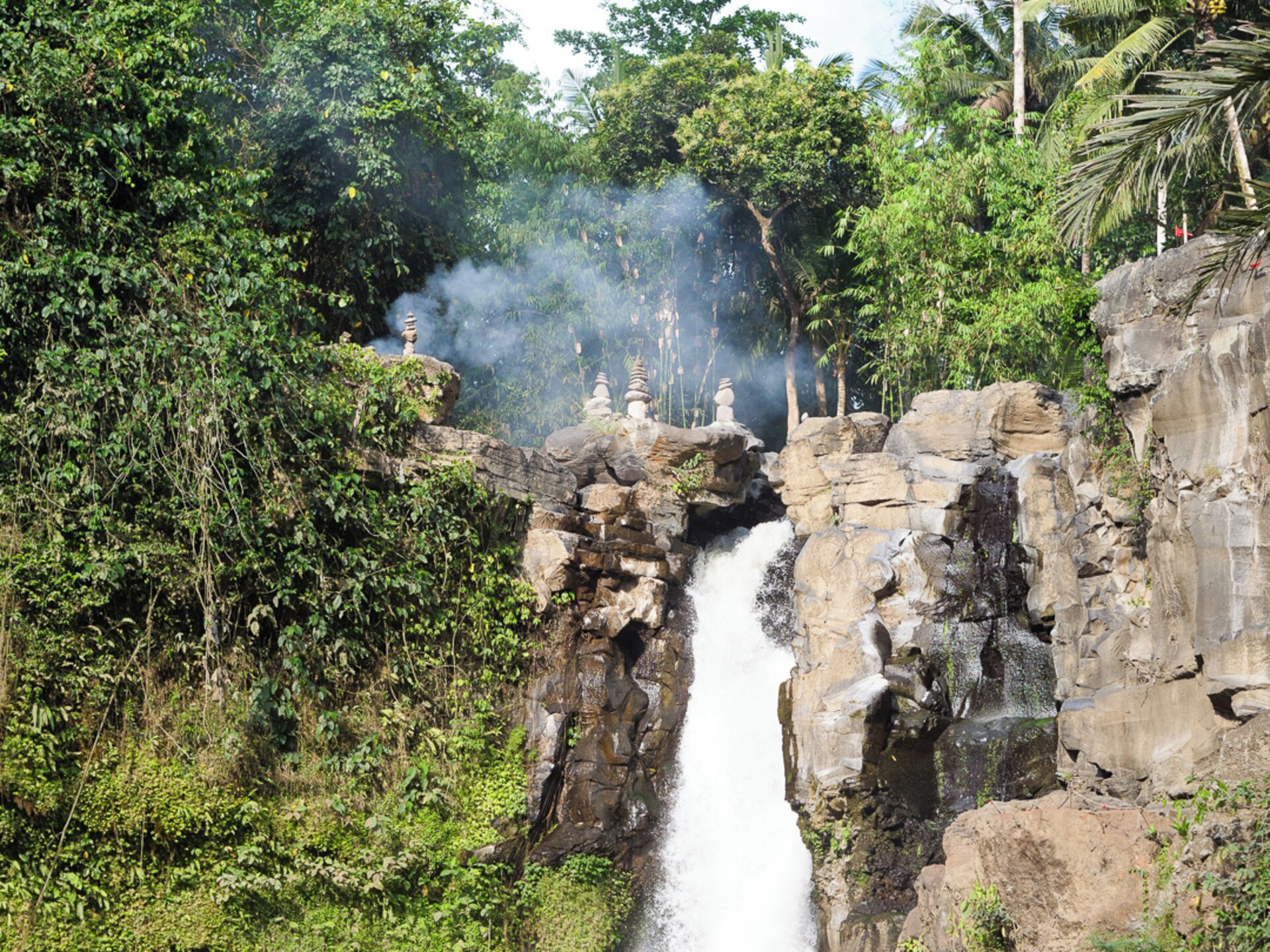 Una cascada se precipita sobre terreno rocoso, con abundante vegetación y construcciones tradicionales.