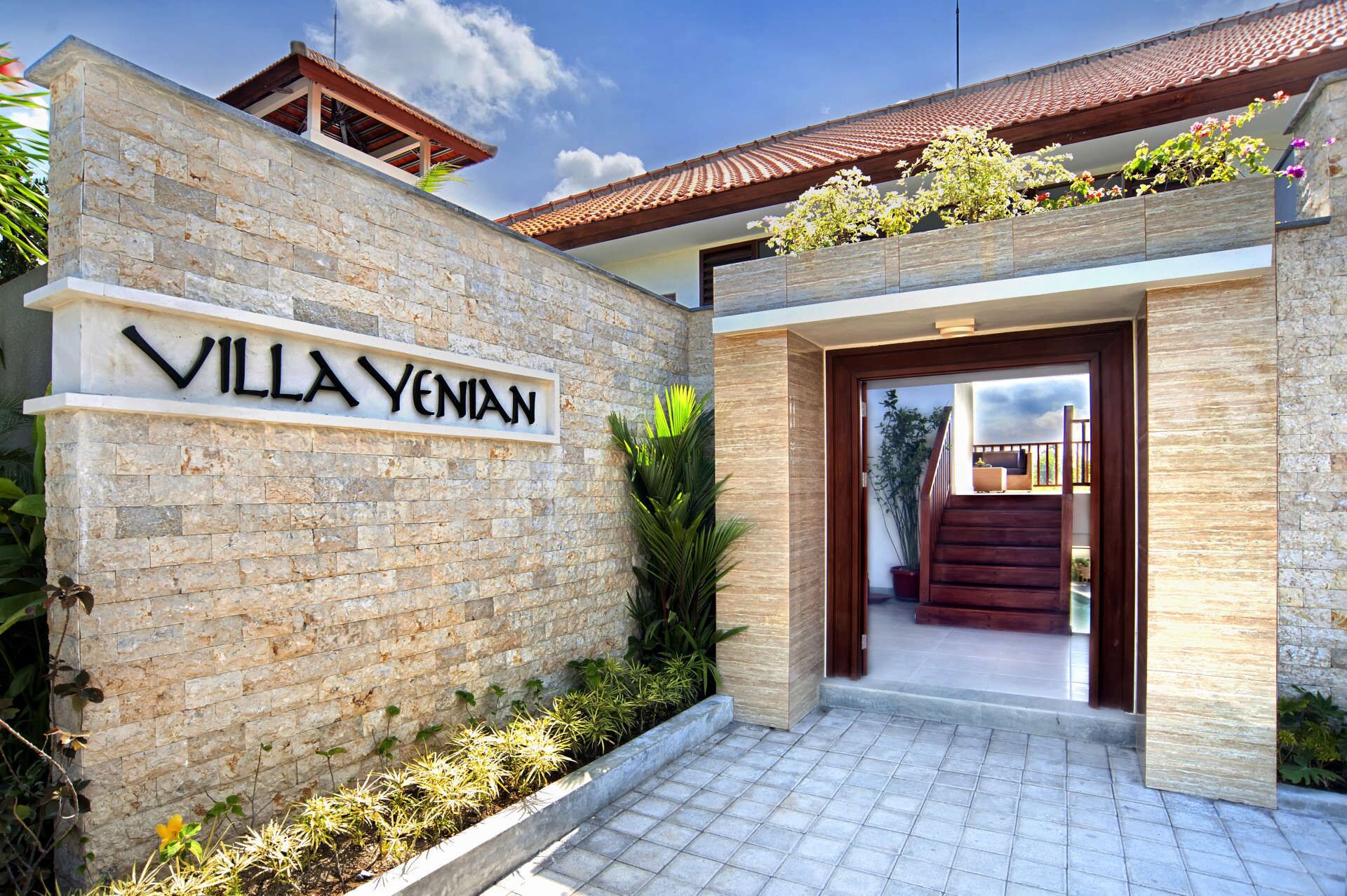 Entrada de una villa con muro de piedra, puerta de madera y un camino empedrado que conduce a un espacio cubierto.