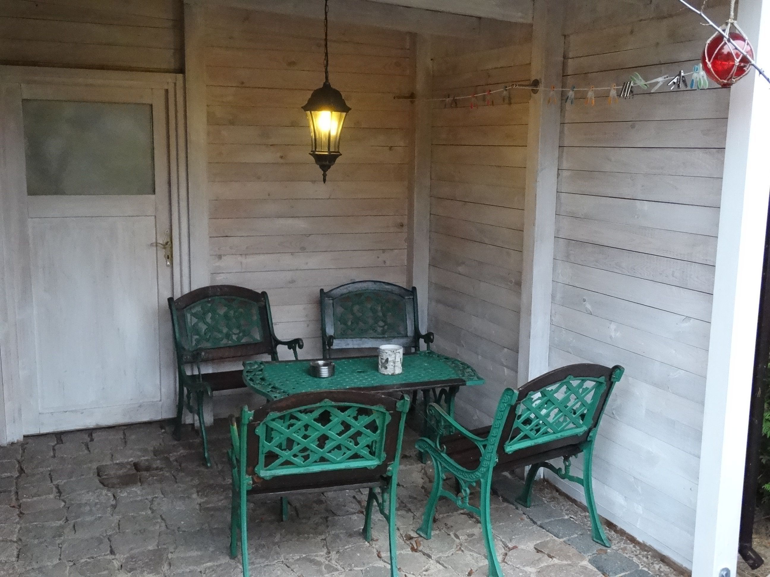 A covered outdoor seating area with a table and four chairs, a hanging lantern, and a wall-mounted drying rack.