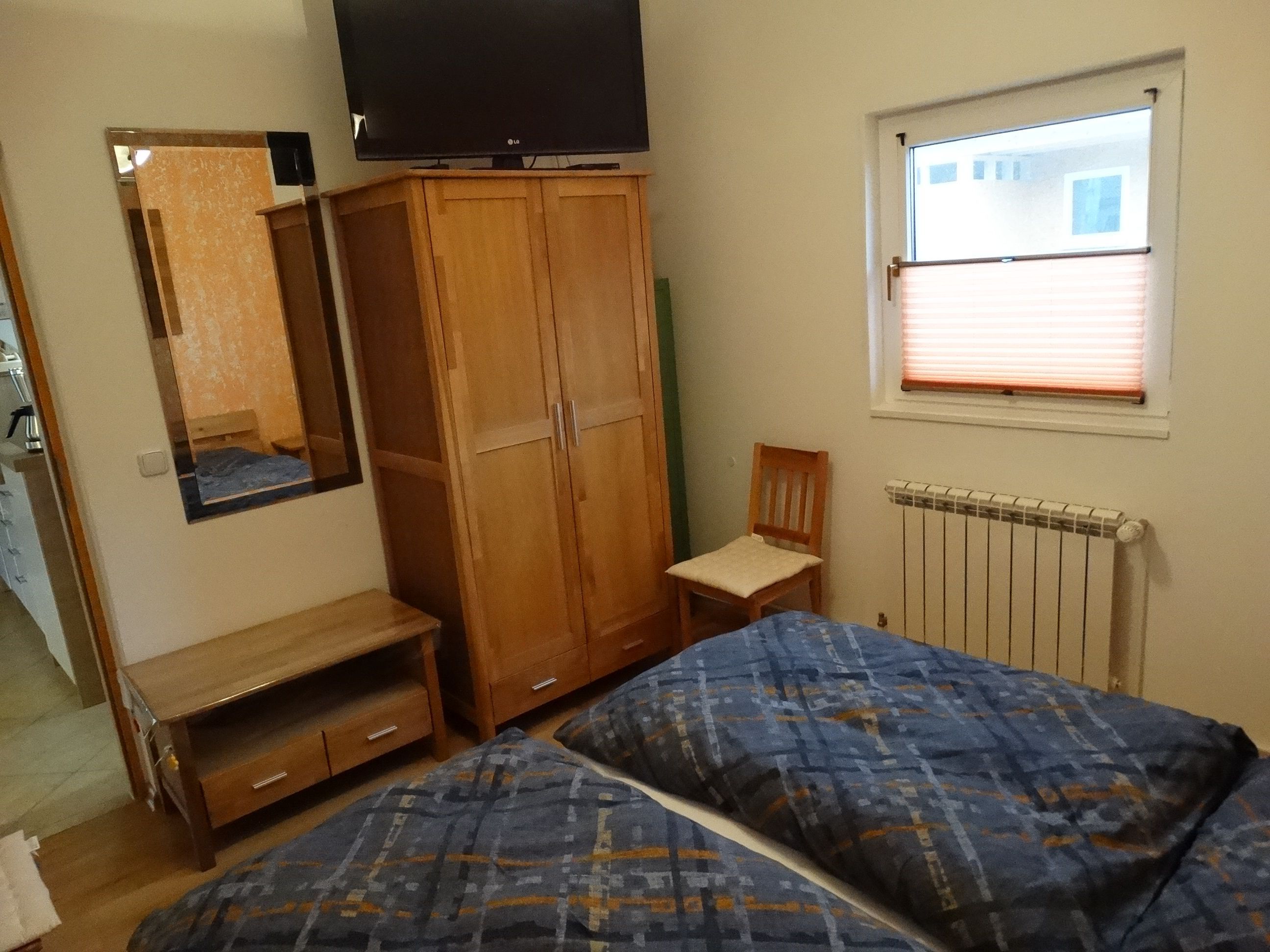 Une chambre à coucher comprenant un lit, une armoire en bois, une chaise, un téléviseur et une fenêtre avec des stores.
