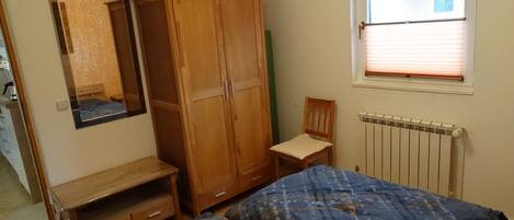 Une chambre à coucher comprenant un lit, une armoire en bois, une chaise, un téléviseur et une fenêtre avec des stores.