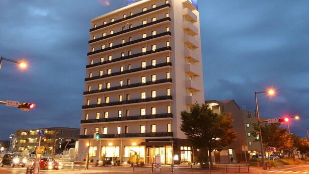 A multi-story hotel with a prominent sign at the top, a ground-level entrance with glass doors, and a parking area in front. - Ab Hotel Kamagori (Gamagori)