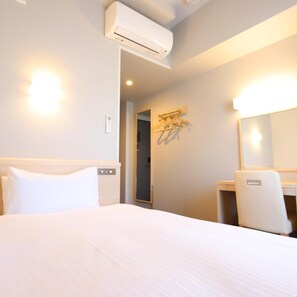 A hotel room with a bed, a desk, a chair, an air conditioning unit, a mirror, and a wall-mounted light fixture.