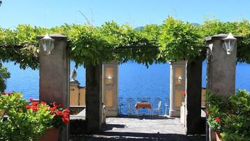 An outdoor patio with a view of the sea, featuring a pergola with climbing plants, a metal railing, and a table with chairs.