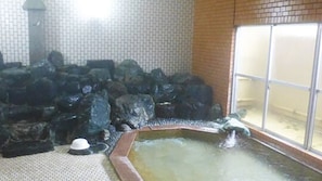 Eine steinene Badewanne, umgeben von Steinen, eine geflieste Wand und ein Fenster mit Rollläden.