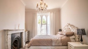 A bedroom with a bed, a nightstand, a lamp, a fireplace, and a window with curtains. - Elegant Georgian 1 Bed Apartment in Herne Bay (Herne Bay)