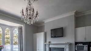 A living room with a fireplace, a chandelier, and a television above it. There is a dining area with a table and chairs, and a kitchen area with cabinets and a microwave. - Elegant Georgian 1 Bed Apartment in Herne Bay (Herne Bay)