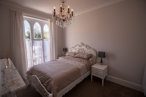 A bedroom with a large bed, a chandelier, a nightstand, and a window with curtains.