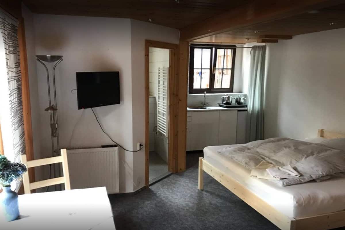 Ein Schlafzimmer mit Bett, Wand-Fernseher, einer kleinen Küchenzeile und einem Fenster mit Vorhängen.