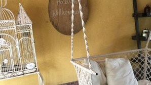 Ein Hängekorbstuhl mit weißen Kissen, ein Metallkübel und ein Holzschild mit der Aufschrift 'Herzlich Willkommen' an einer gelben Wand.