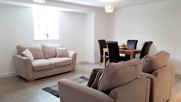 A modern living room with a beige sofa, a dining table with chairs, and a large window with natural light. - Riverside 5 (Maidstone)
