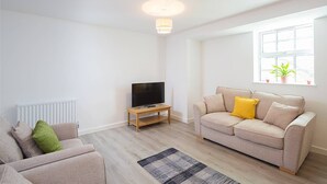 A modern living room with a beige sofa, a television on a wooden stand, a radiator, and a window with a plant.