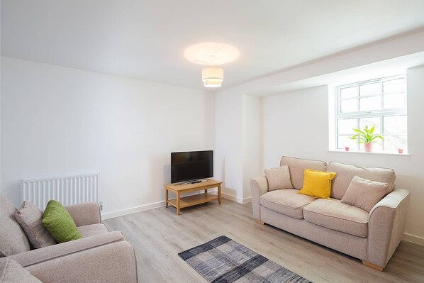 A modern living room with a beige sofa, a television on a wooden stand, a radiator, and a window with a plant.
