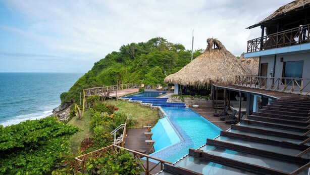 Un resort con piscina, terraza de madera y construcciones con techo de paja.