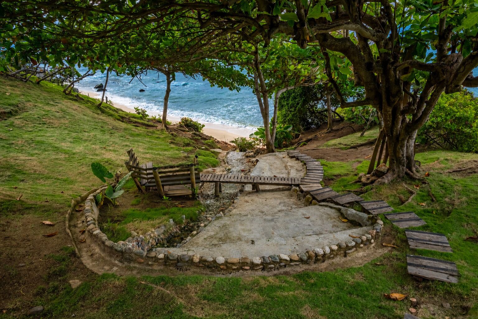 Un patio de piedra circular con un banco de madera y un pequeño puente, rodeado de árboles y con vistas a una playa.