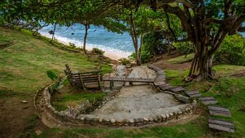 Un patio de piedra circular con un banco de madera y un pequeño puente, rodeado de árboles y con vistas a una playa.