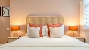 Ein ordentlich bezogenes Bett mit weißen und pfirsichfarbenen Kissen, einem Korbhaupt und zwei Nachttischlampen auf Nachttischen.