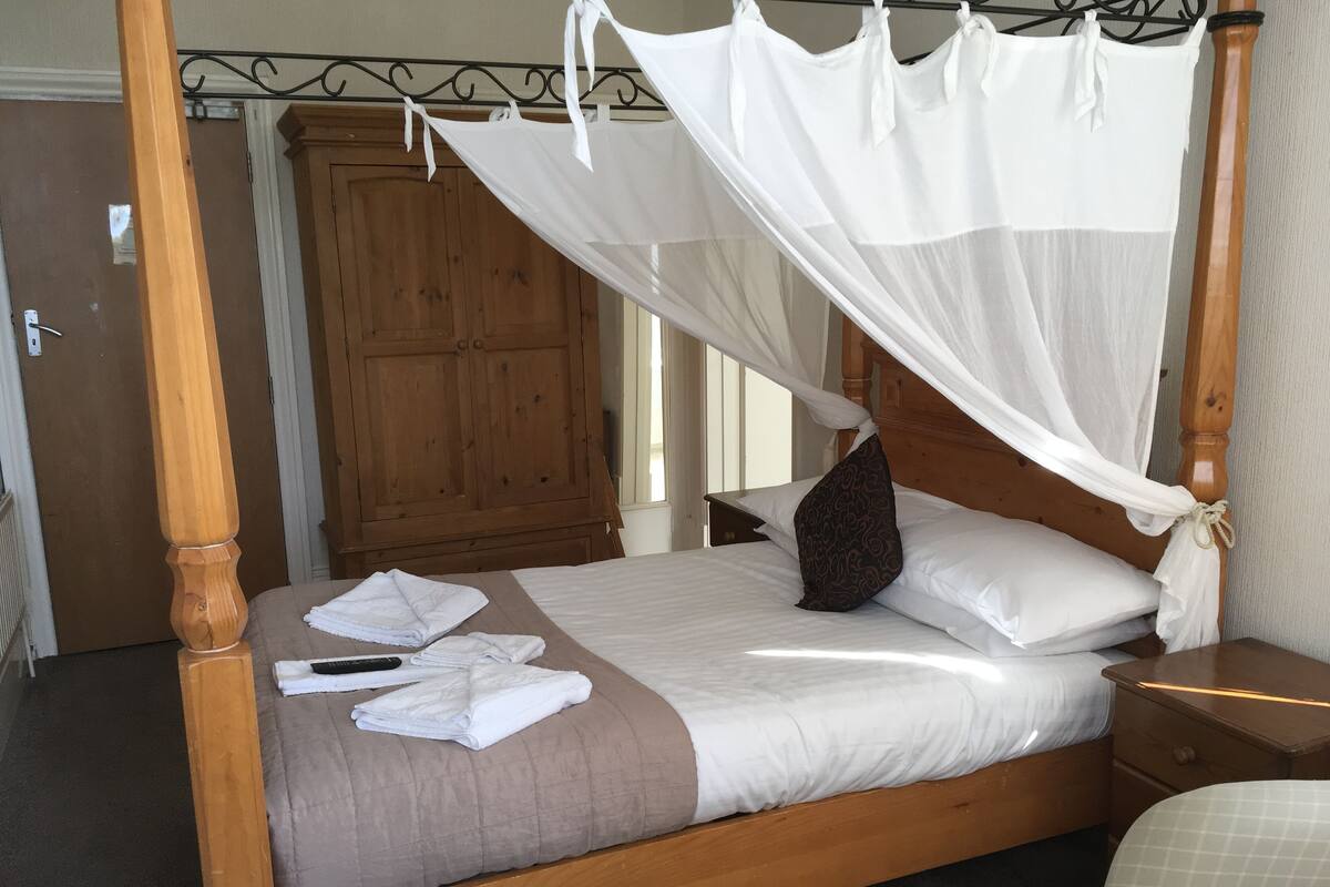 Uma cama de dossel com cortina, um guarda-roupa de madeira e uma mesa de cabeceira com abajur.