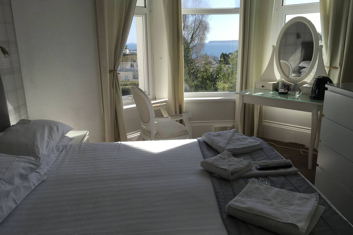 Uma cama bem arrumada com almofadas, uma cadeira, uma escrivaninha com espelho e vista para o mar pela janela.