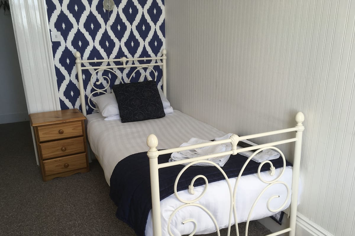 Cama bem arrumada com estrutura de metal branca, colcha azul escura e travesseiro preto. Um criado-mudo de madeira com gavetas fica à esquerda da cama. O quarto tem papel de parede estampado e uma parede lisa à direita.