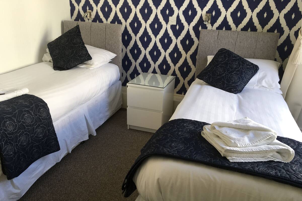 Quarto de hotel com duas camas de solteiro, cada uma com colcha estampada escura e roupa de cama branca. Há uma pequena mesa de cabeceira com abajur e uma gaveta branca. As paredes possuem papel de parede com padrão geométrico.