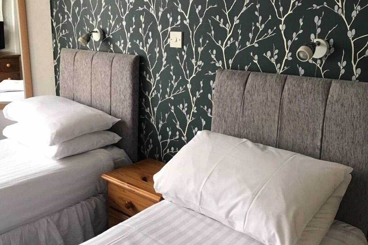 Quarto com duas camas, uma mesinha de cabeceira e uma parede com padrão de folhagem.
