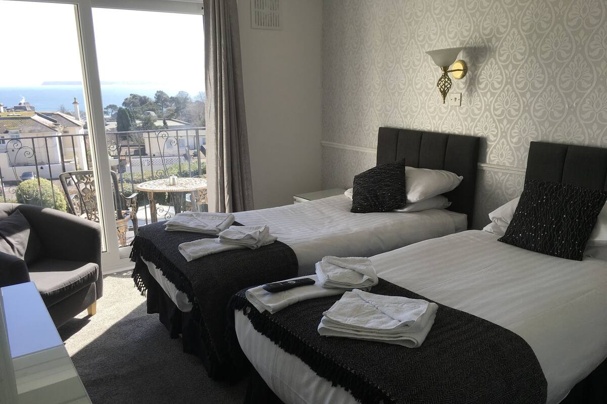 Quarto de hotel com duas camas, uma cadeira e uma mesa. Vista da cidade pela janela.
