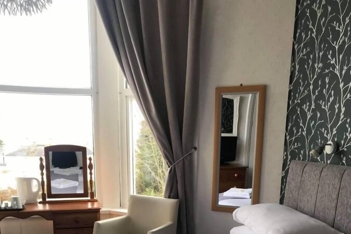Quarto de hotel com duas camas, uma escrivaninha com espelho, uma cadeira e uma janela com cortinas.
