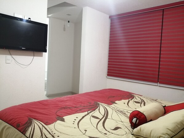 Un dormitorio con una cama, un televisor montado en la pared y una ventana con persianas rojas.