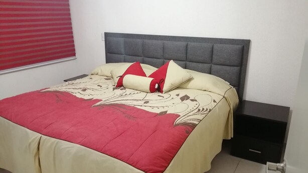 Cama bem arrumada com colcha vermelha e bege, cabeceira estofada cinza e uma pequena mesa de cabeceira com gaveta.