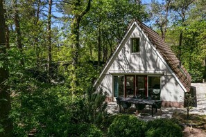 A small, white wooden cabin with a sloped roof, surrounded by dense forest.