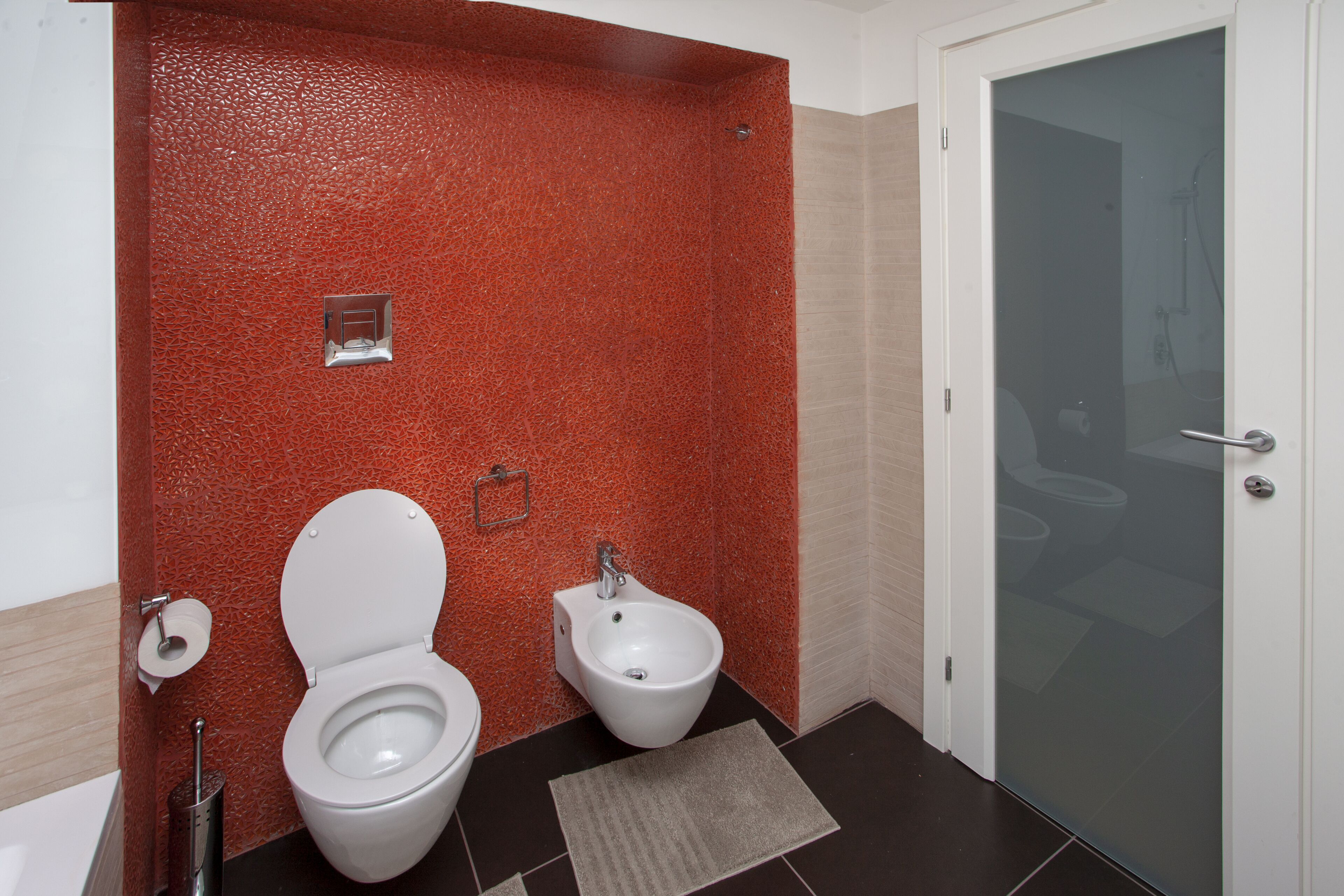 Un baño con pared roja con textura, un inodoro blanco, un bidé y una puerta de ducha de vidrio.