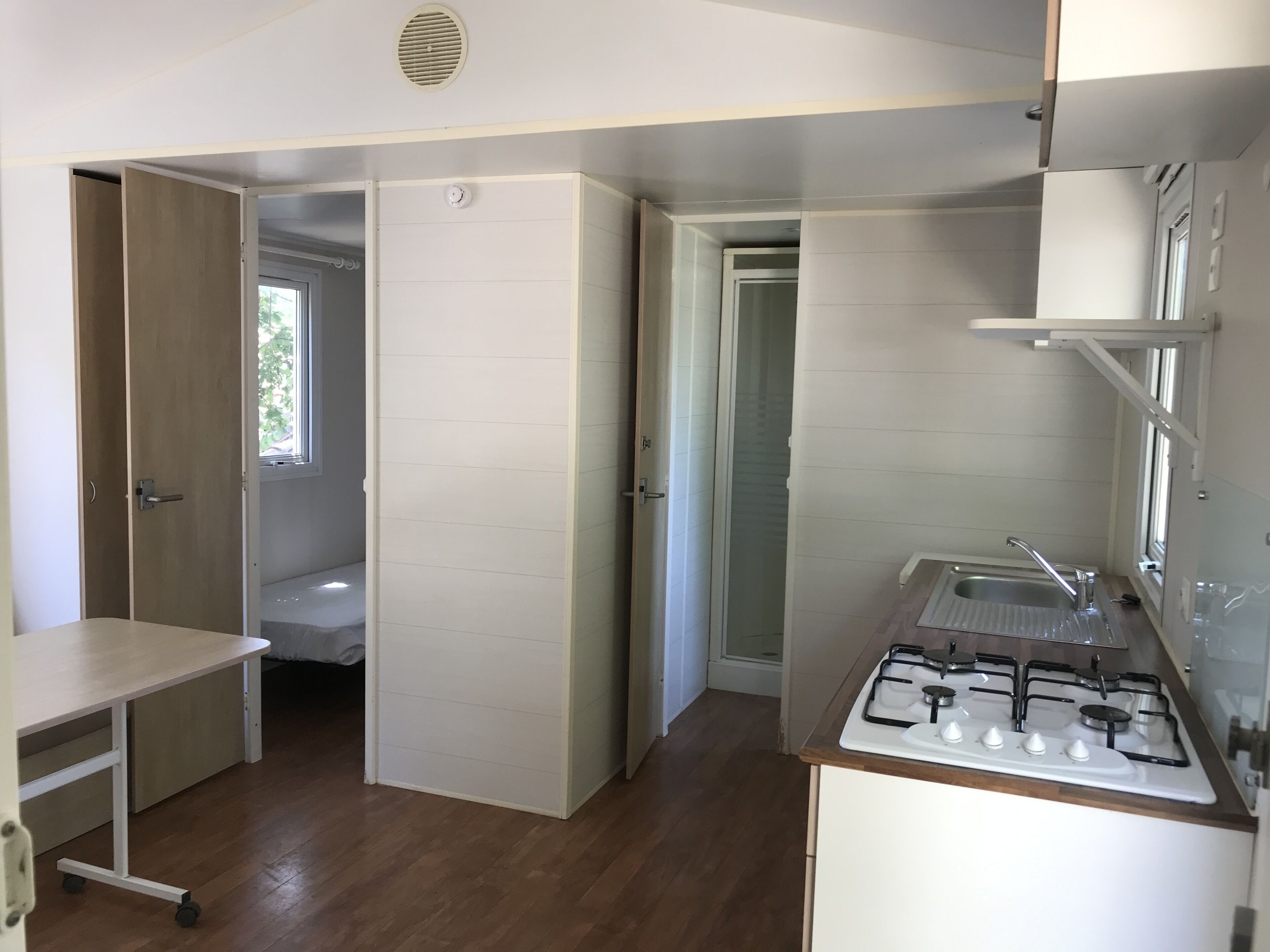 Eine kleine Küche mit Gasherd, Spüle und einer kompakten Arbeitsfläche. Eine Tür führt ins Badezimmer, ein Fenster sorgt für natürliches Licht.